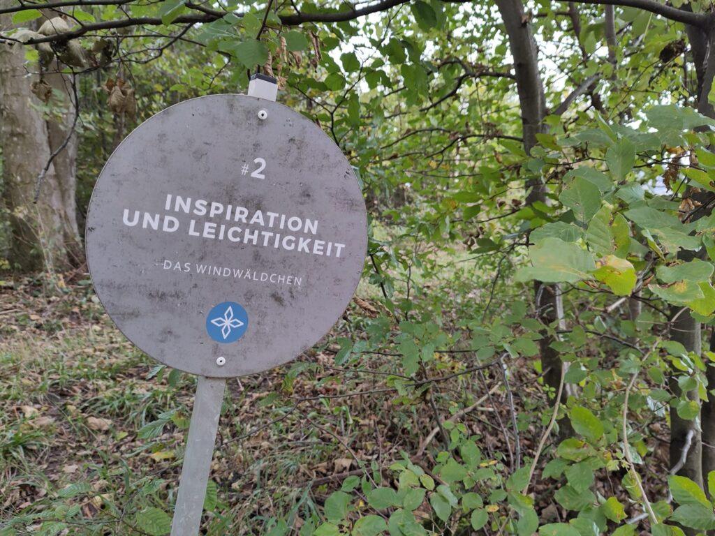 Rundes Schild mit der Aufschrift '#2 Inspiration und Leichtigkeit Das Windwäldchen' in einem Wald mit grünen Blättern