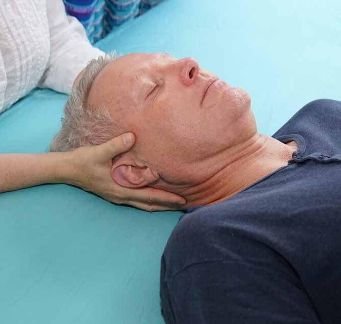Person liegt auf Rücken auf Behandlungsliege, eine andere Person hält sanft den Kopf zur craniosacralen Therapie
